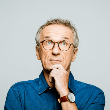Portrait of elderly man wearing blue denim shirt and glasses looking up with hand on chin. Thoughtful senior entrepreneur, studio shot against grey background.